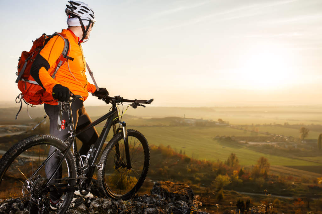Man with the bike standing on a rock. spring nature. Cyclist in orange jacket, helmet and with red backpack. Healthy traveling in the countryside. Landscape with field, hill and rocks. Background of sky.